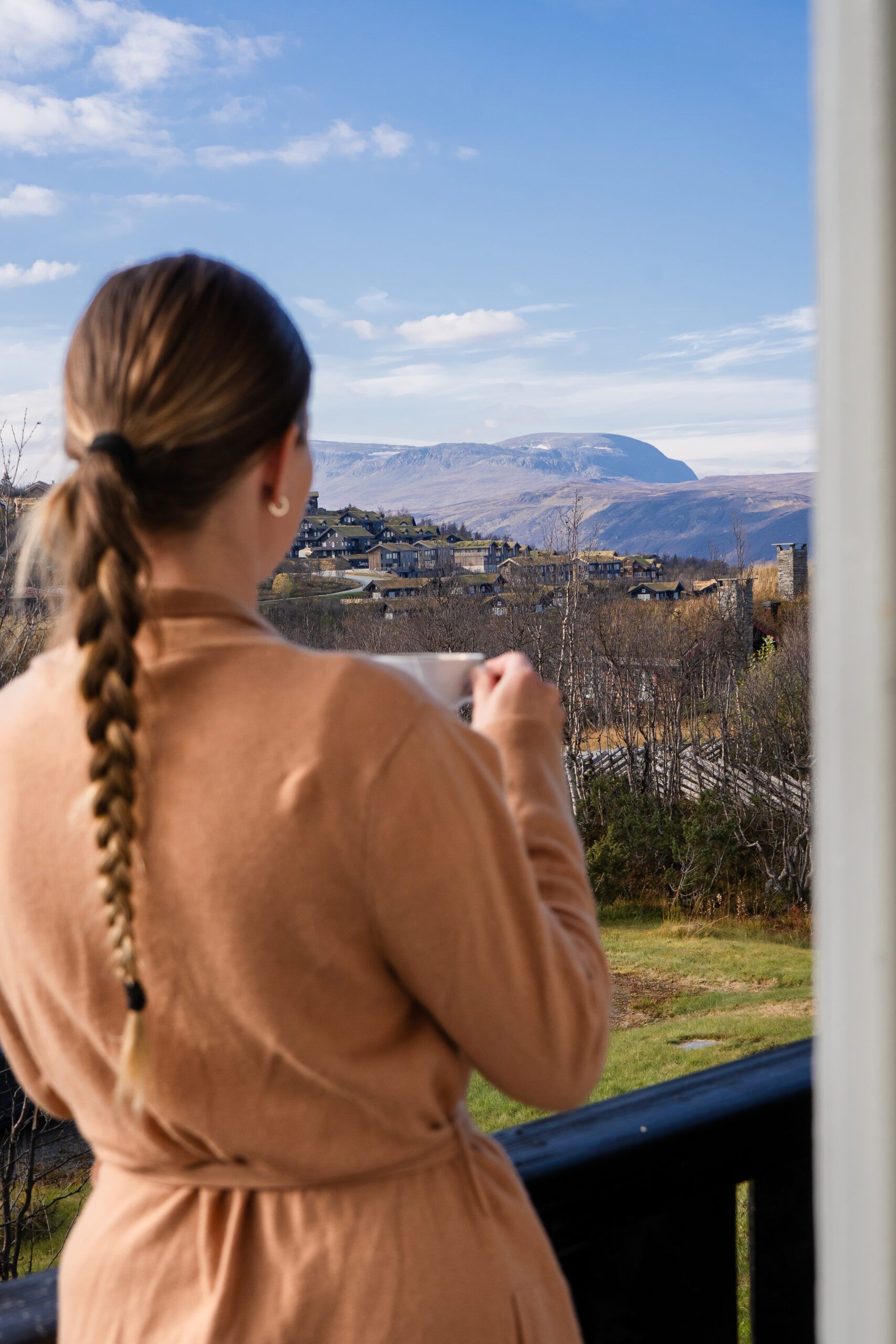 Tovebu - Kikut utsikt Kvinne står utendørs på en balkong og holder en kopp i hånden mens hun speider utover fjellandskap. Foto