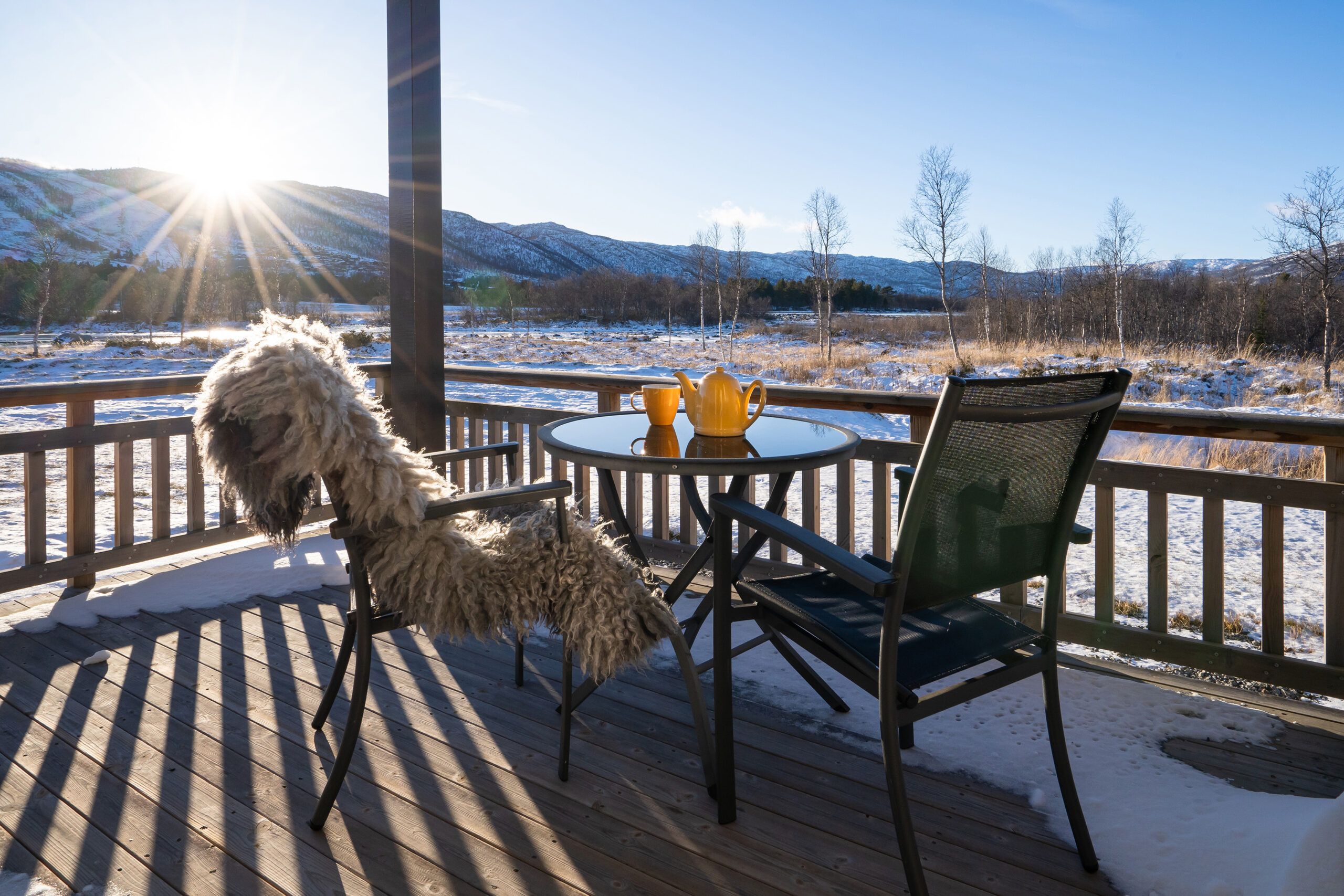 Veranda med bord og stoler. En tekanne og en kopp står på bordet, og det ligger et saueskinn i den ene stolen. Solen er på vei bak fjellet, og landskapet er dekket av snø. Foto