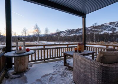 Veranda med utemøbler og lykter. Utsikt over skibakke. Snø ligger både på plattingen og i omgivelsene. Kveldslys med rosa tendenser på himmelen. Foto